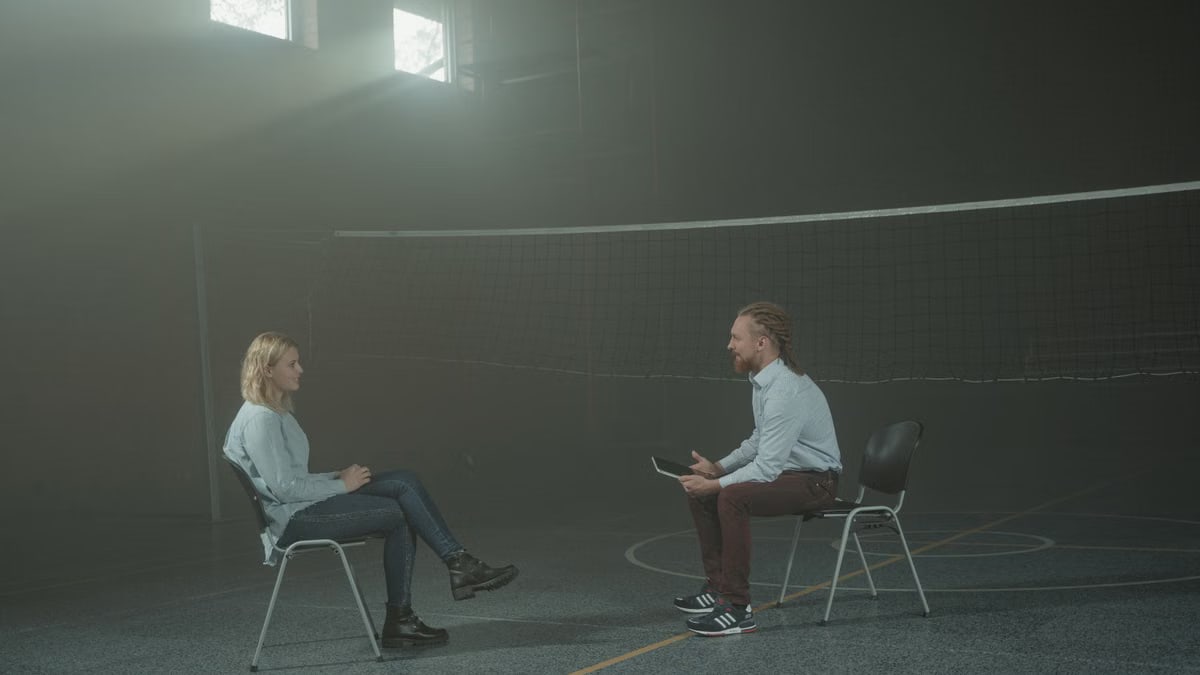 A man and a woman engage in a therapy session in a smoky indoor setting, sitting apart in a large room.
